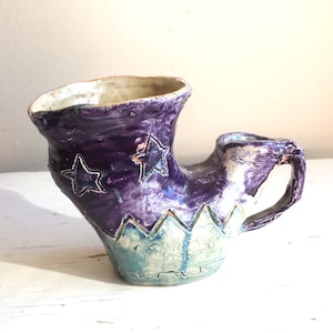 Könnte beinhalten: Eine lila und blaue Keramiktasse mit einem Sternendesign. Die Tasse hat eine einzigartige Form mit einem Griff, der seitlich herausragt.