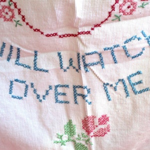 May include: A pink fabric with blue embroidery that reads "WILL WATCH OVER ME". The fabric is decorated with red and green embroidered flowers.