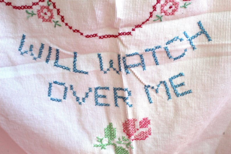 May include: A pink fabric with blue embroidery that reads "WILL WATCH OVER ME". The fabric is decorated with red and green embroidered flowers.