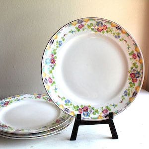 May include: Set of four white ceramic dinner plates with a floral border in shades of pink, yellow, blue, and green. The plates have a gold rim and are stacked on a black stand.