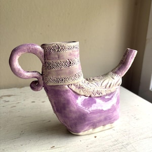 May include: A ceramic pitcher with a glossy purple glaze and a white base. The vessel features a curved handle, a long spout, and textured bands around the neck and body with geometric patterns including hexagons and leaf-like impressions.