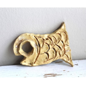 Puede incluir: Una escultura de pez de cerámica hecha a mano en color beige claro. El pez tiene detalles tallados para representar escamas y aletas, y una gran boca abierta. La escultura se exhibe sobre una superficie blanca.