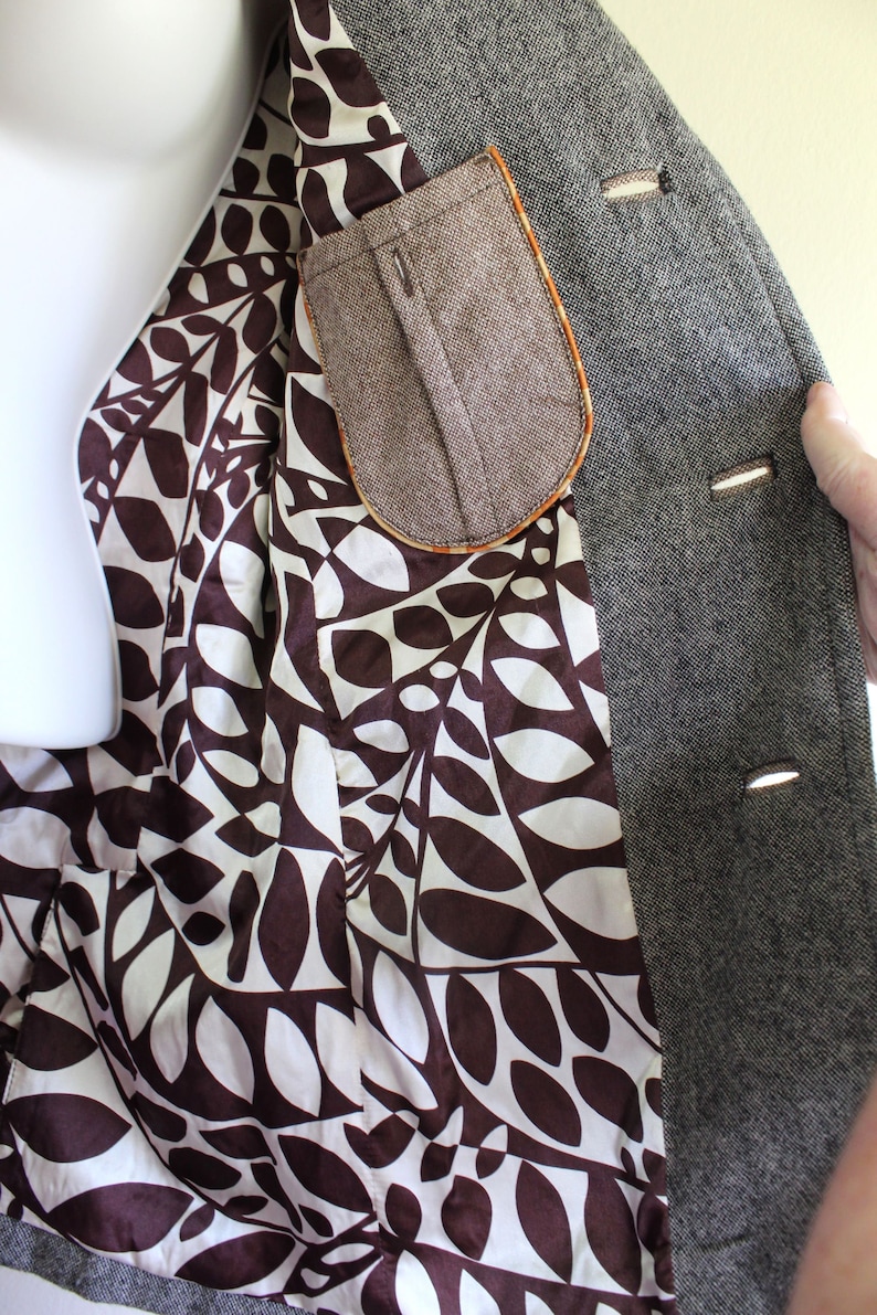May include: A grey tweed blazer with a brown leaf patterned lining and a brown tweed pocket. The blazer has three buttons on the front.