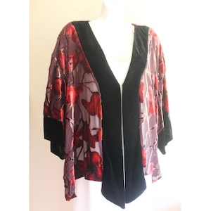 May include: A sheer, open-front jacket with a floral pattern in shades of red and purple. The jacket features black velvet trim along the edges and sleeves. The garment is displayed on a mannequin, showcasing its design and drape.