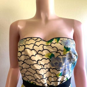 May include: A strapless bodice featuring a cream-colored fabric with a repeating black scalloped pattern and a floral print of blue flowers with green leaves. The neckline is trimmed with black piping, and a solid black band sits at the waistline.
