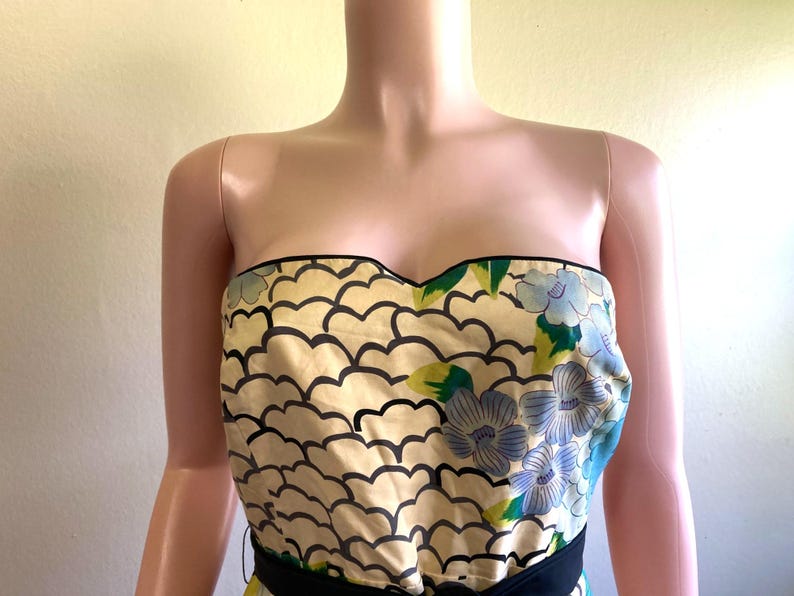 May include: A strapless bodice featuring a cream-colored fabric with a repeating black scalloped pattern and a floral print of blue flowers with green leaves. The neckline is trimmed with black piping, and a solid black band sits at the waistline.