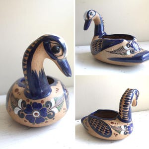 Può includere: Fioriera a forma di cigno in ceramica dipinta a mano con base beige e dettagli blu. Il cigno presenta un collo lungo e curvo e un motivo floreale dettagliato. La fioriera misura circa 15 cm di lunghezza e 10 cm di altezza.