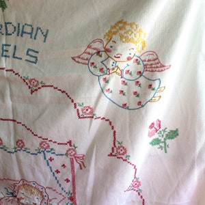 May include: White cotton embroidered tablecloth with pink, blue, and green floral designs. The tablecloth features a cross-stitch design of an angel with the text "Guardian Angels".