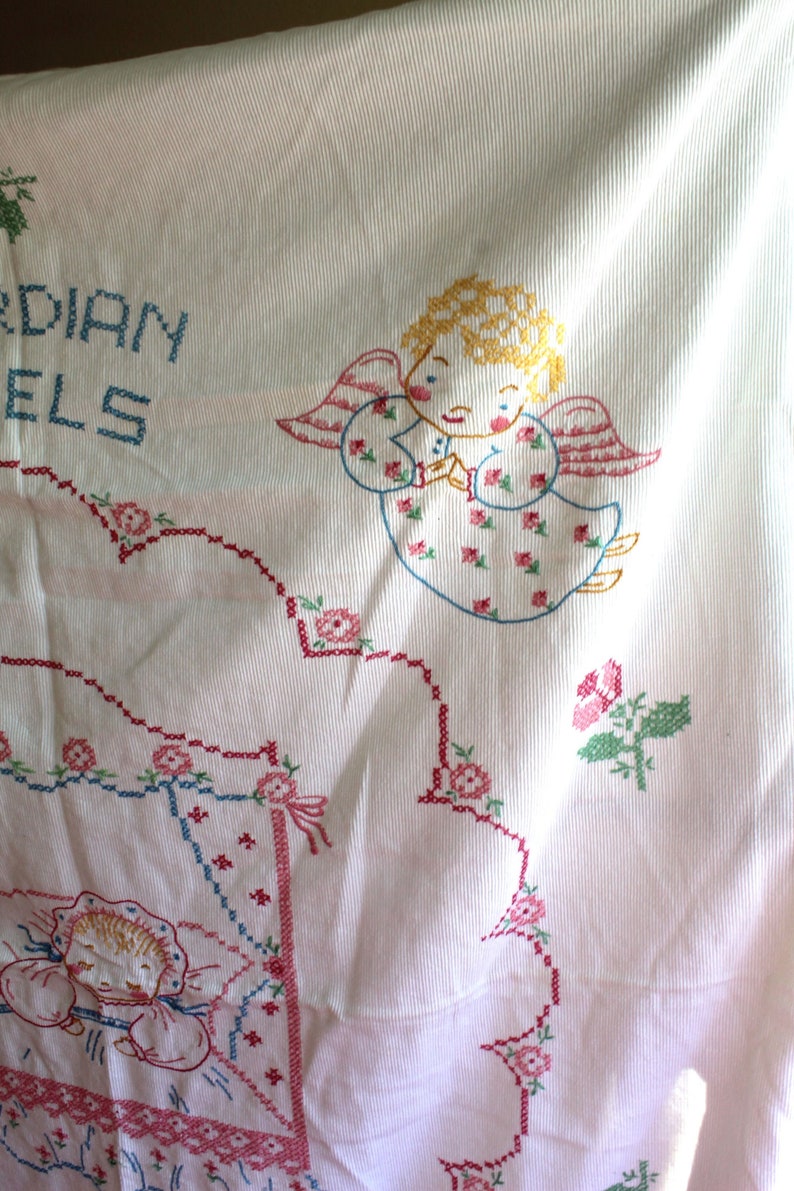 May include: White cotton embroidered tablecloth with pink, blue, and green floral designs. The tablecloth features a cross-stitch design of an angel with the text "Guardian Angels".