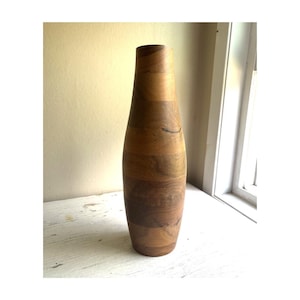 Könnte beinhalten: Eine hohe, zylindrische Vase aus braunen Holzsegmenten mit sichtbarer natürlicher Maserung. Die Vase hat eine glatte, konische Form, die sich zur oberen Öffnung hin leicht verjüngt und zum Boden hin breiter wird.