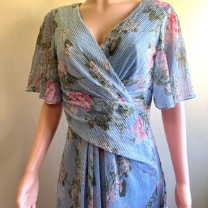 Op de afbeelding: Een lichtblauwe jurk met een bloemenpatroon. De jurk heeft een V-hals, korte fladdermouwen en een wikkelbustier. De stof heeft een subtiele geplooide textuur en het bloemmotief bevat roze en groene accenten.