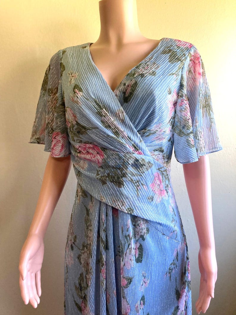Op de afbeelding: Een lichtblauwe jurk met een bloemenpatroon. De jurk heeft een V-hals, korte fladdermouwen en een wikkelbustier. De stof heeft een subtiele geplooide textuur en het bloemmotief bevat roze en groene accenten.