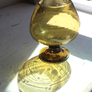 Può includere: Un calice vintage in vetro color ambra con una coppa arrotondata e un gambo corto. Il vetro ha una leggera consistenza e proietta un'ombra dettagliata sulla superficie bianca. Il calice è posizionato vicino a una finestra.
