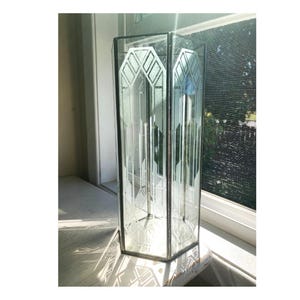 Könnte beinhalten: Eine hohe, klare Glasvase mit geätzten geometrischen Mustern. Die Vase hat eine rechteckige Form mit abgeschrägten Kanten und einem dekorativen Muster. Sie steht auf einer weißen Fensterbank, wobei Sonnenlicht Schatten wirft. Die Vase ist etwa 30 cm hoch.