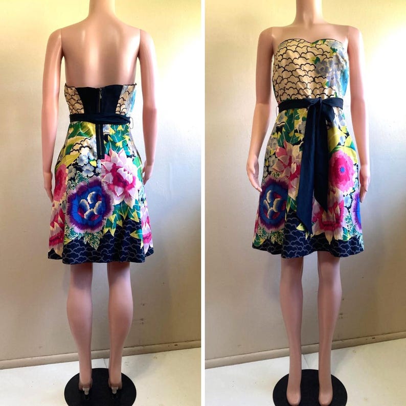 May include: A strapless knee-length dress featuring a vibrant floral print with large pink, blue, and green flowers on a cream background. The dress includes a solid black waist tie and a black zipper closure on the back, with a dark wave pattern at the hem.