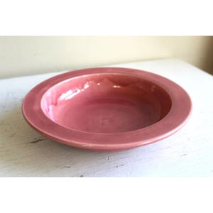 May include: A round, shallow, ceramic bowl in a muted rose color. The bowl has a wide, flat rim and a smooth, glossy finish. The interior of the bowl is slightly lighter in color than the rim. The bowl is sitting on a white surface.