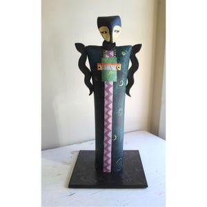 May include: A tall, abstract sculpture of a person with a black head and a teal, purple, and green patterned body. The sculpture is standing on a black square base.