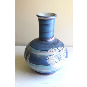 Może przedstawiać: Ręcznie wykonany ceramiczny wazon o okrągłym korpusie i wąskiej szyi. Wazon ma niebiesko-białą kolorystykę z teksturowanym, malowanym wzorem. Wzór zawiera białe, kropkowane kształty kwiatowe i poziome niebieskie paski.
