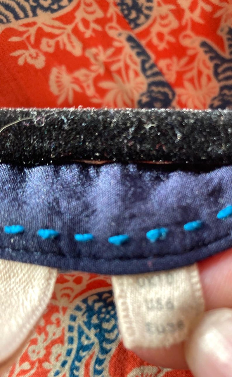 May include: Close-up of a textile with a navy blue fabric edge, accented with bright blue stitching. The background features a vibrant red and white patterned fabric. A tag with text is visible.