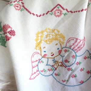 May include: White cotton fabric with embroidered angel with blonde hair and pink wings. The angel is wearing a blue dress with pink and green flowers. The fabric has a red and green floral border.