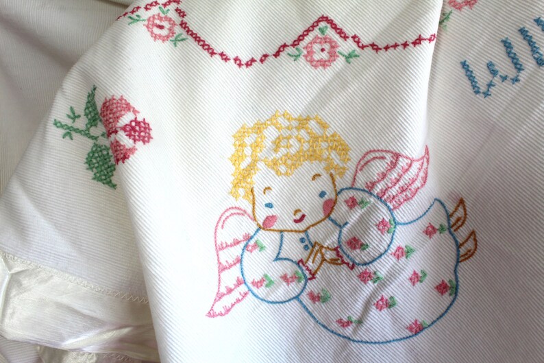 May include: White cotton fabric with embroidered angel with blonde hair and pink wings. The angel is wearing a blue dress with pink and green flowers. The fabric has a red and green floral border.