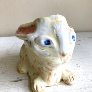 Könnte beinhalten: Keramik-Hasenfigur mit blauen Augen, rosa Ohrspitzen und einer hellgelb-braunen, melierten Oberfläche. Der Hase sitzt mit ausgestreckten Pfoten. Die Figur hat ein skurriles, handgemachtes Aussehen.