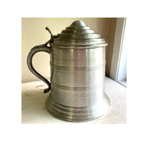 May include: A vintage silver-colored beer stein with a hinged lid and a curved handle. The stein has a cylindrical body with horizontal bands and a flared base. The lid is conical with concentric rings. The handle is ornate.