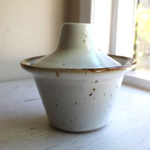 Puede incluir: Un pequeño recipiente de cerámica blanquecina con tapa. El recipiente tiene una base acampanada y un borde redondeado con un borde marrón. La tapa es cónica y la cerámica tiene un aspecto moteado.
