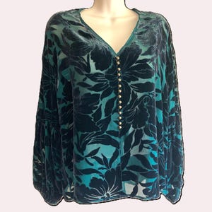 May include: A teal and navy floral print blouse with a V-neckline and long sleeves. The blouse features a button-down front with gold-colored buttons. The fabric has a velvet-like texture and a semi-sheer appearance.