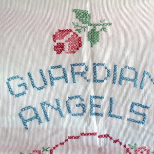 May include: White fabric with blue embroidery that reads "GUARDIAN ANGELS". A red and green embroidered rose is above the text.