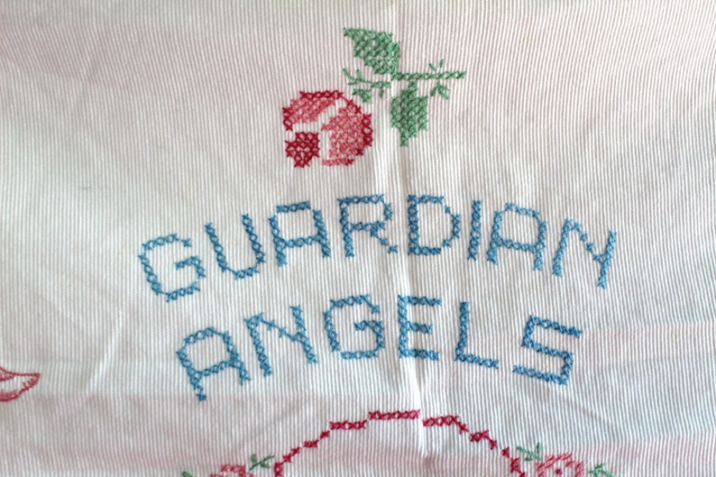 May include: White fabric with blue embroidery that reads "GUARDIAN ANGELS". A red and green embroidered rose is above the text.