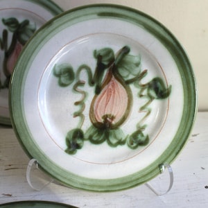 May include: White ceramic plate with a green border and a hand-painted design. The design features a pink fruit with green leaves and vines. The plate is displayed on a clear stand.