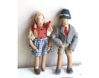 May include: Two vintage cloth dolls, a girl and a boy, stand side-by-side. The girl doll wears a red top with black polka dots and a blue and white plaid skirt. The boy doll wears a gray jacket, shorts, and a green hat. Both dolls have painted faces.