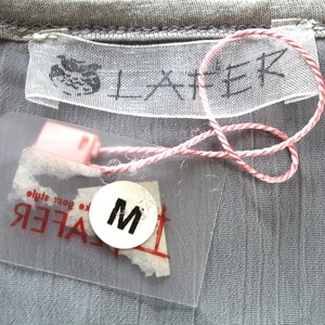 May include: A white tag with the word "LAFER" printed in black. A clear plastic tag with the word "LAFER" printed in black and a size "M" printed in black on a white circle. The tags are attached to a grey fabric with a red and white string.