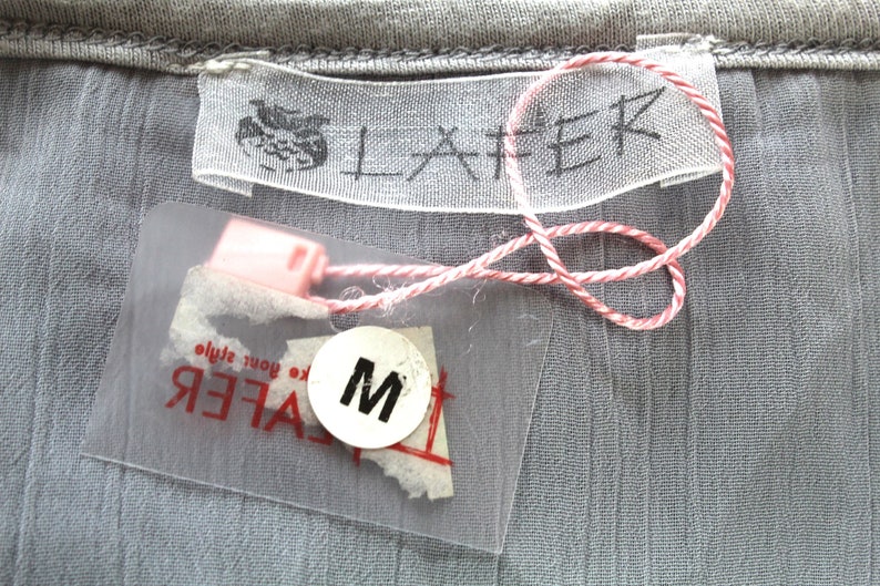 May include: A white tag with the word "LAFER" printed in black. A clear plastic tag with the word "LAFER" printed in black and a size "M" printed in black on a white circle. The tags are attached to a grey fabric with a red and white string.