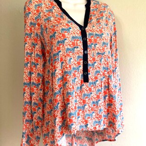 May include: A long-sleeved blouse with a vibrant pattern of blue zebras on a coral background. The blouse features a V-neckline with a navy trim and a button-down front. The fabric has a slightly textured appearance.