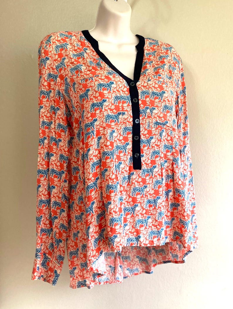 May include: A long-sleeved blouse with a vibrant pattern of blue zebras on a coral background. The blouse features a V-neckline with a navy trim and a button-down front. The fabric has a slightly textured appearance.