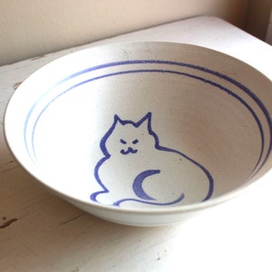 May include: White ceramic bowl with a blue cat design. The cat is sitting with its tail curled around its legs. The bowl has a blue rim.