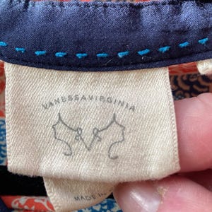 May include: Close-up of a cream-colored fabric tag with the text "VANESSAVIRGINIA" in black. Below the text are two stylized drawings of women's profiles. The tag is sewn onto a navy blue fabric with blue stitching.