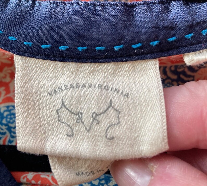May include: Close-up of a cream-colored fabric tag with the text "VANESSAVIRGINIA" in black. Below the text are two stylized drawings of women's profiles. The tag is sewn onto a navy blue fabric with blue stitching.