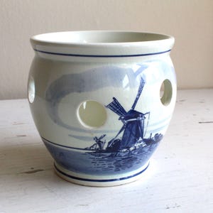 May include: A white ceramic bulb planter with a rounded body and four circular cutouts. The exterior features a blue transferware-style illustration of a Dutch windmill landscape, accented by thin blue horizontal lines around the rim and base.