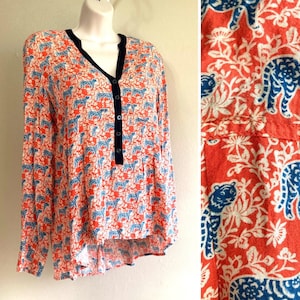 May include: A long-sleeved blouse with a vibrant pattern of blue tigers and white floral designs on a coral background. The blouse features a V-neckline with a navy trim and a button-down front.