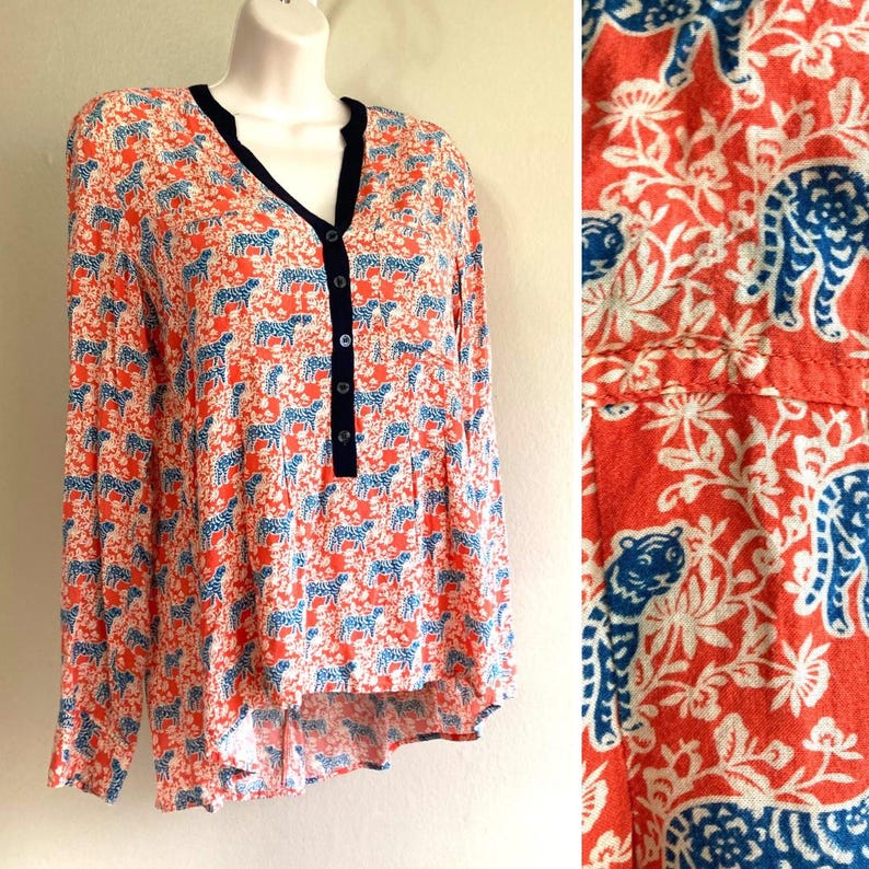 May include: A long-sleeved blouse with a vibrant pattern of blue tigers and white floral designs on a coral background. The blouse features a V-neckline with a navy trim and a button-down front.