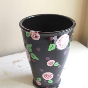 Puede incluir: Un jarrón de cerámica negro con un diseño floral. El jarrón está decorado con rosas pintadas de rosa y verde y puntos.