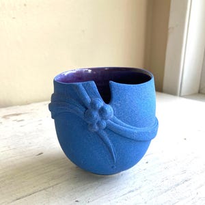 Pode incluir: Um vaso de cerâmica azul texturizado com um design único. O vaso apresenta uma fita esculpida e enfeites florais na frente. O interior é roxo escuro, contrastando com o exterior. O vaso tem uma seção recortada na parte superior.