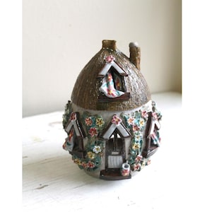 Może przedstawiać: Mały ceramiczny domek z dachem ze słomy, ozdobiony kwiatami i oknami. Domek jest pomalowany w odcieniach brązu, zieleni i bieli.