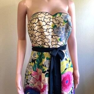 May include: A strapless dress featuring a sweetheart neckline and a black waist sash. The bodice displays a cream-colored pattern of black scalloped lines, while the skirt showcases a vibrant floral print in shades of pink, blue, green, and yellow.