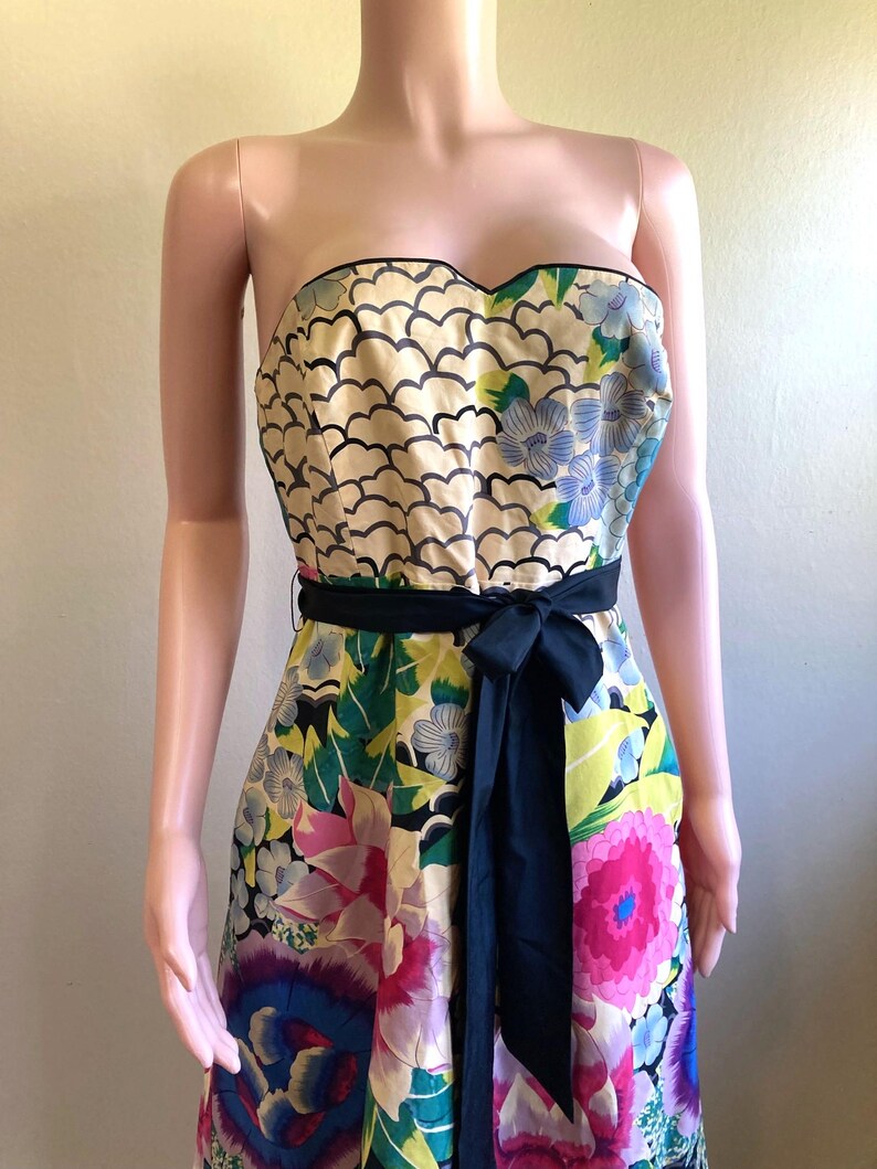 May include: A strapless dress featuring a sweetheart neckline and a black waist sash. The bodice displays a cream-colored pattern of black scalloped lines, while the skirt showcases a vibrant floral print in shades of pink, blue, green, and yellow.