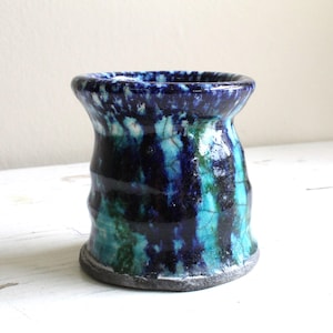 May include: A handmade ceramic vase with a unique, textured surface. The vase features a blend of deep blue, teal, and black colors, creating a marbled effect. The rim is slightly flared, and the base is a darker shade. A decorative piece for the home.