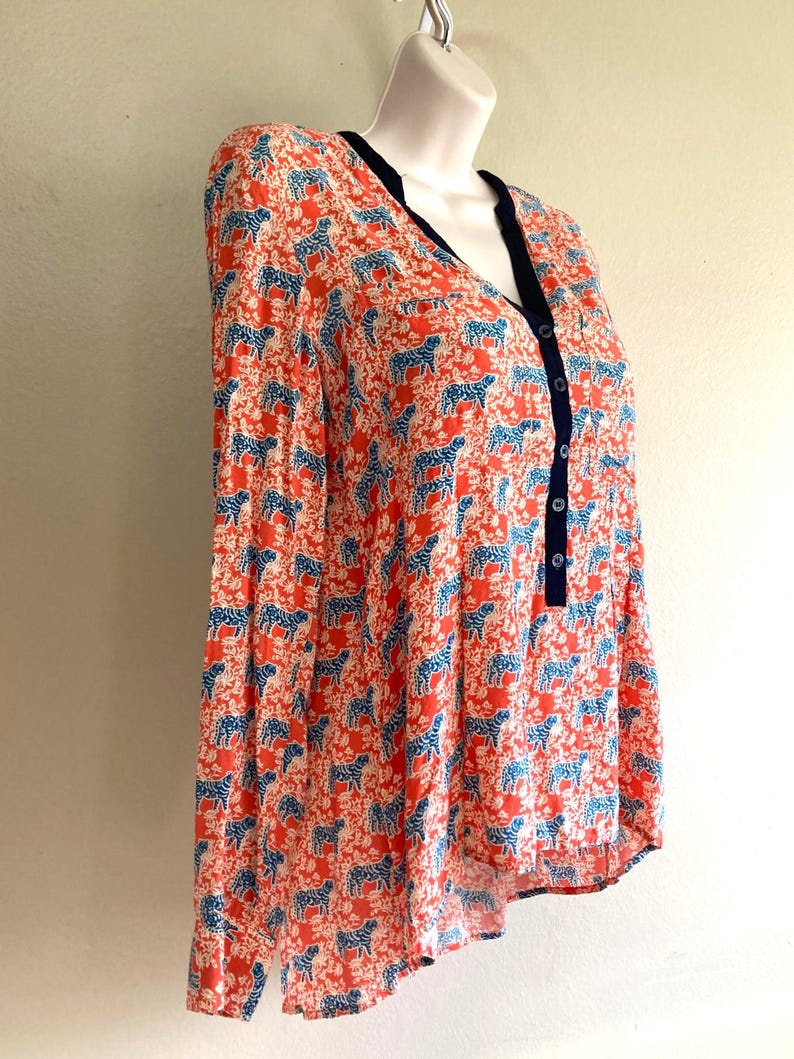 May include: A long-sleeved blouse with a vibrant pattern of blue animal figures on a coral background. The blouse features a V-neckline with navy trim and a button-down front. The fabric appears lightweight and suitable for warmer weather.